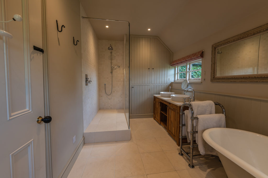 Spacious bathroom with a walk-in shower on the left, featuring a glass partition. Twin sinks with round mirrors and a wooden vanity are against the far wall. A freestanding bathtub with towels is on the right. Indulge in luxury travel vibes as neutral tones adorn both walls and floor. David Pearson Travel