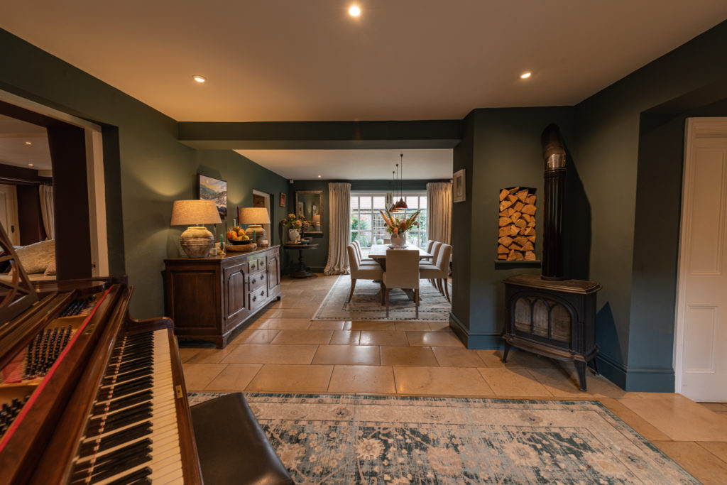 A cozy living room exuding luxury travel vibes, featuring a piano on the left, a dining area with an elegant table and chairs in the back, and a wood-burning stove on the right. Warm lighting illuminates the green walls adorned with artwork and sophisticated decorative items. David Pearson Travel