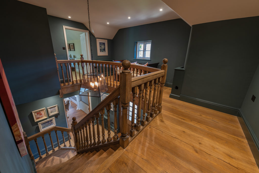 A wooden staircase with an ornate railing leads to the upper floor, where dark green walls and wooden flooring evoke the charm of luxury travel. A chandelier hangs above, illuminating framed art that decorates the walls. A small window lets in light, and a doorway at the top leads to another room. David Pearson Travel