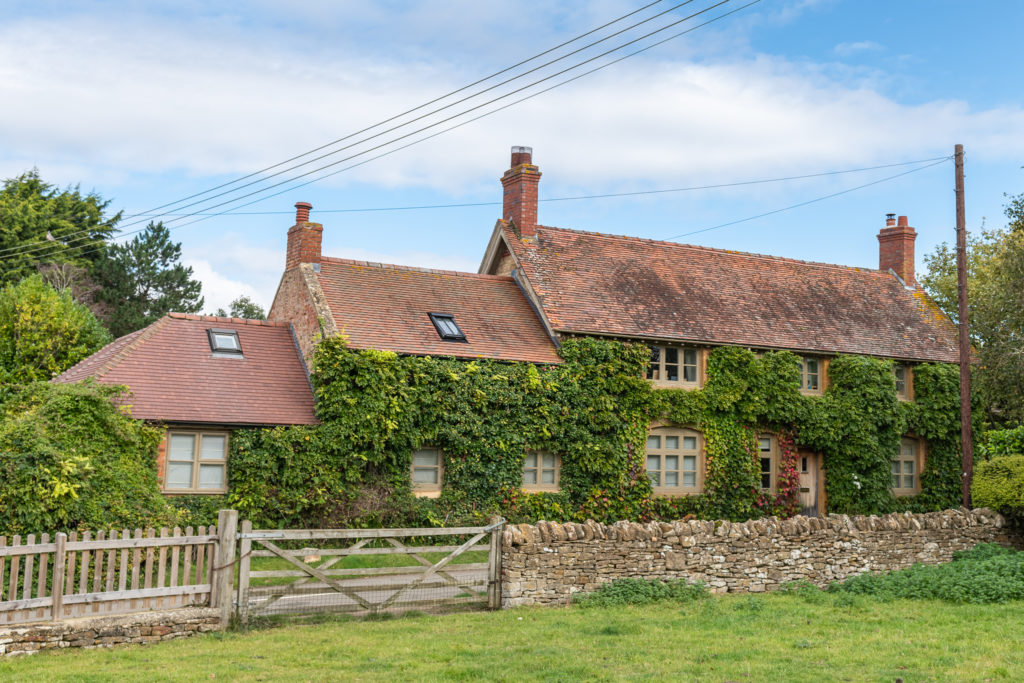 Nestled in a lush countryside, this charming cottage exudes luxury travel vibes with its ivy-covered brick walls, multiple chimneys, and red-tiled roofs. A stone fence encircles the enchanting retreat, all under a partly cloudy sky that enhances its allure. David Pearson Travel