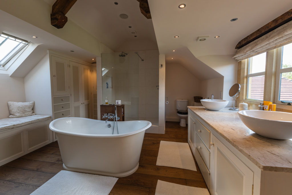 Luxurious bathroom reminiscent of a luxury travel retreat, featuring a freestanding bathtub, wooden flooring, large walk-in shower, and double sinks. Beamed ceilings and a skylight enhance natural light over the elegant decor, white cabinets, and the cozy bench by the window. David Pearson Travel