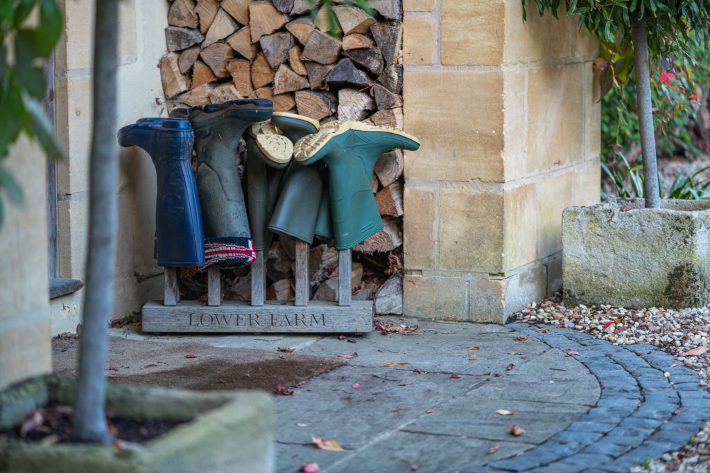 Four pairs of boots are neatly arranged on a wooden rack labeled LOWER FARM, nestled against a brick wall stacked with firewood. This charming outdoor scene, perfect for those seeking a touch of rustic luxury travel, is complemented by nearby trees and stone planters. David Pearson Travel