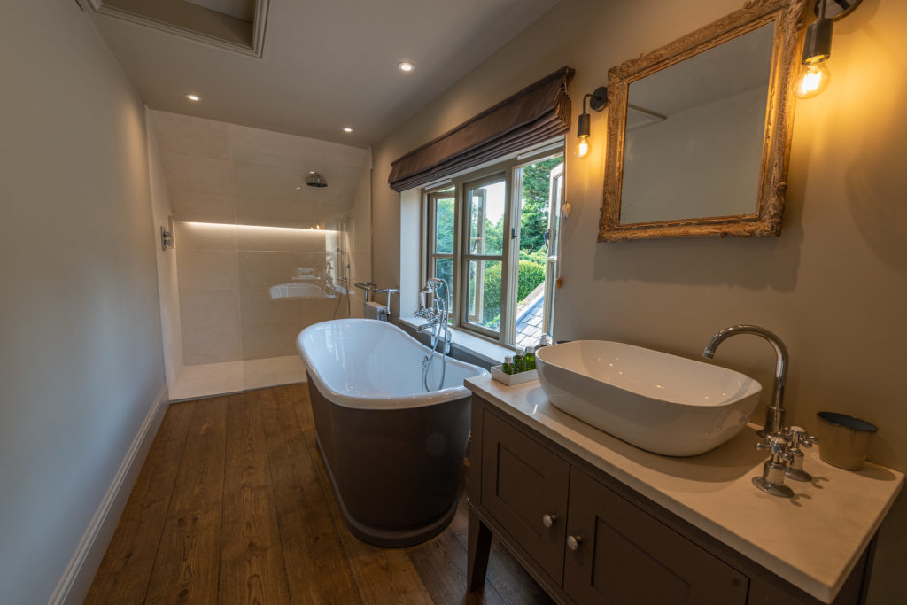 A modern bathroom exuding a luxury travel vibe features a freestanding bathtub, a vessel sink on a wooden vanity, and a large mirror. A walk-in shower with glass paneling complements the wooden flooring. A window reveals lush greenery outside, while warm lighting enhances the lavish space. David Pearson Travel