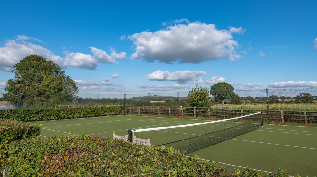 A tennis court, epitomizing luxury travel, is surrounded by lush green bushes and a wooden fence under a vibrant blue sky with scattered clouds. Trees and open fields stretch into the background, meeting a distant hill on the horizon. David Pearson Travel