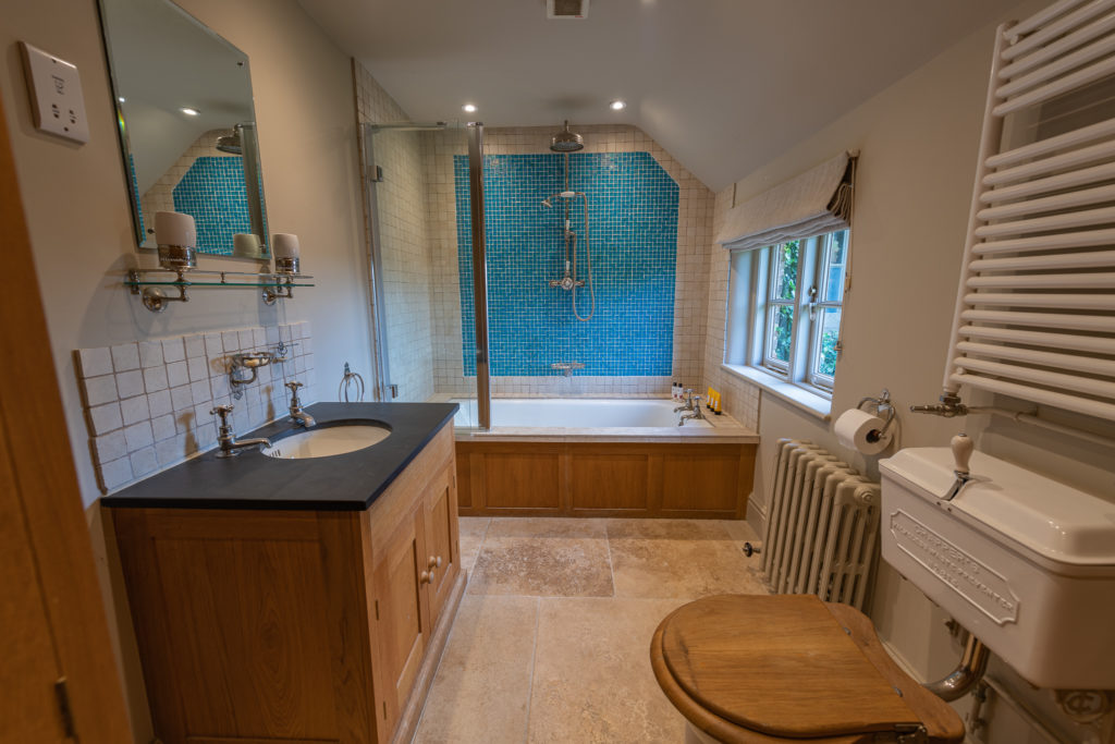 Experience luxury in this bathroom oasis, featuring a wooden vanity with a black countertop and sleek sink, complemented by a mirror and a toilet with a wooden seat. Relax in the bathtub with its glass enclosure against blue tiled walls, and enjoy the comfort of a heated towel rail. David Pearson Travel