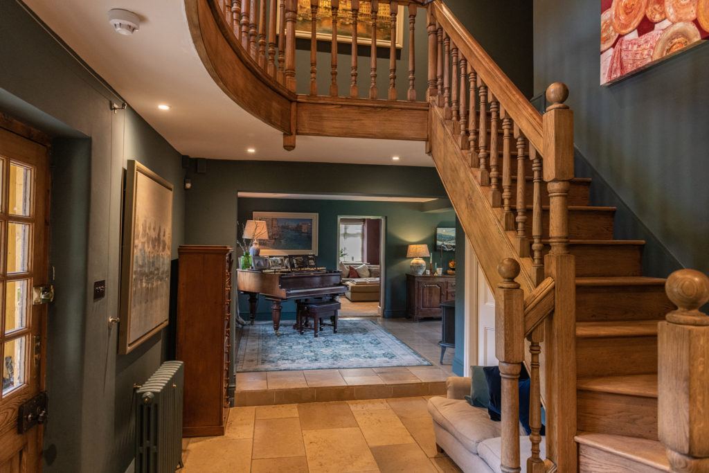 A cozy hallway with wooden stairs leads to a warmly lit room, embodying the essence of luxury travel. A grand piano stands on a blue rug next to large paintings and a sofa. The deep green walls complement the earthy tones of the wooden accents and beige tiled floor. David Pearson Travel