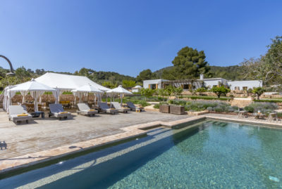 Experience the essence of luxury travel in this exquisite outdoor setting, where a clear swimming pool glistens alongside lounge chairs and white canopy tents on a wooden deck. Behind lies an elegant house nestled amid lush greenery and trees, all under a bright blue sky. David Pearson Travel