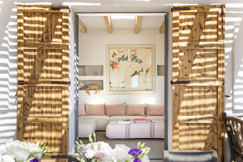 A cozy indoor seating area with a cushioned sofa and decorative pillows evokes the essence of luxury travel. Natural light streams through open wooden shutters, casting striped shadows. A colorful art piece hangs on the wall, while flowers blur in the foreground. David Pearson Travel