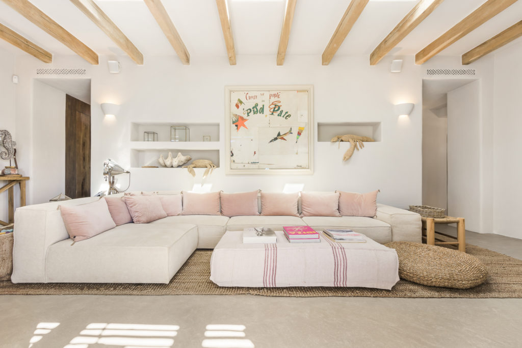 A bright living room with a large white sectional sofa adorned with pink cushions awaits like a luxury travel escape. A woven rug and round pouf sit on a light floor, while framed artwork graces the wall above. Wooden beams line the ceiling, and recessed shelves hold curated decor items. David Pearson Travel