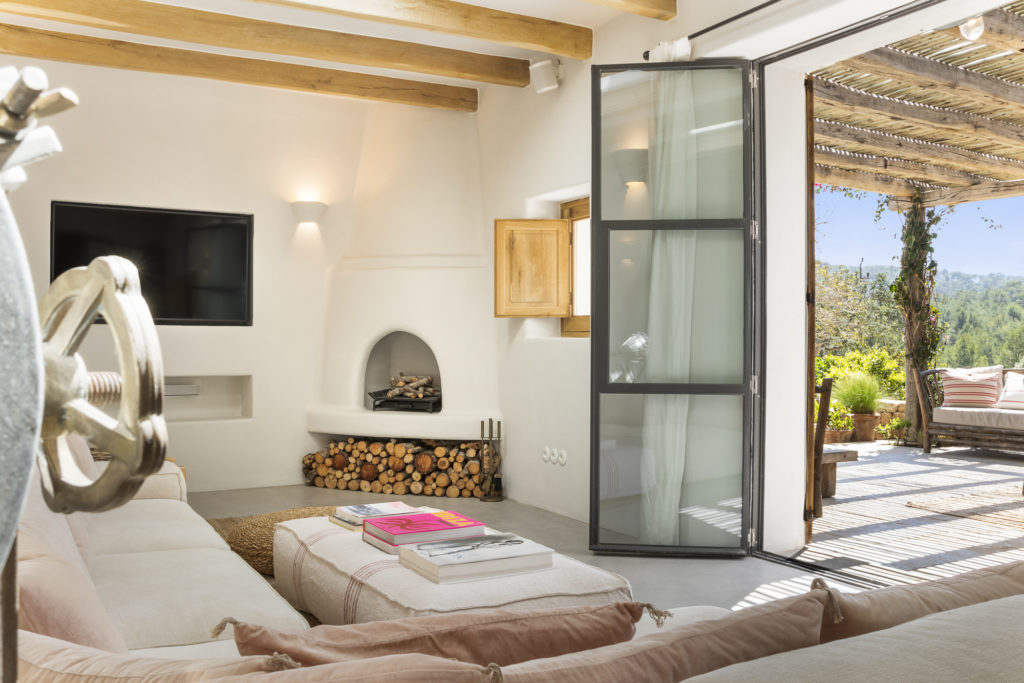 The bright living room exudes luxury with its wood-beamed ceiling, beige sofa, and TV. A small fireplace with stacked logs nestles against a white wall. Large glass doors reveal a sunny patio with lush greenery and distant views, crafting a cozy, inviting space ideal for luxury travel relaxation. David Pearson Travel