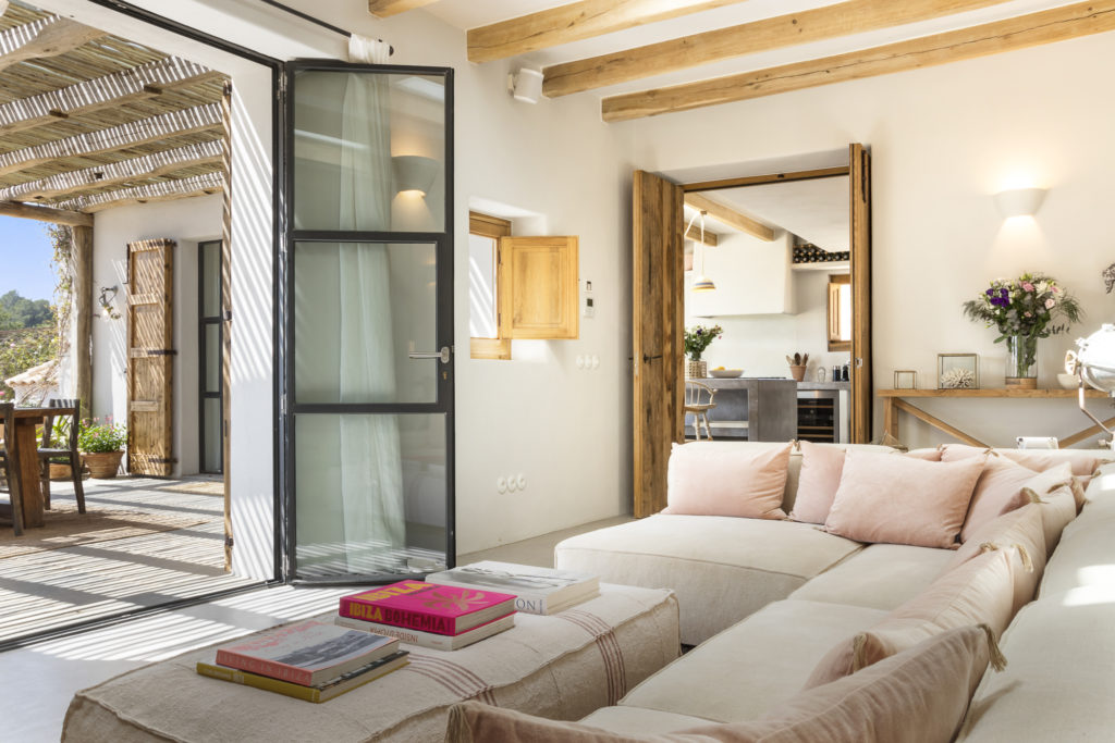 A cozy living room with a large beige sectional sofa adorned with pink cushions exudes luxury travel vibes. Open wooden doors lead to a sunlit terrace, reminiscent of an exotic getaway. Wooden beams and light streaming through a glass door complete this inviting space, with the kitchen visible in the background. David Pearson Travel