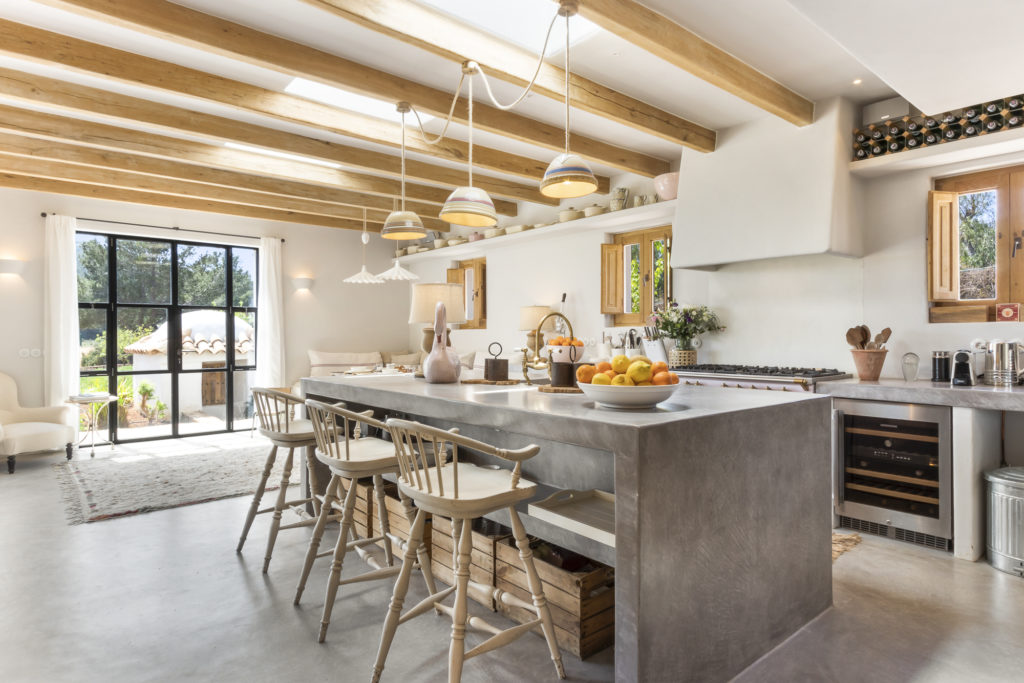 A spacious kitchen evokes the elegance of luxury travel with a long gray island and wooden bar stools. The ceiling features wooden beams and hanging lights, while large windows flood the space with natural light. Modern appliances complement the inviting fruit bowl and decorative items. David Pearson Travel