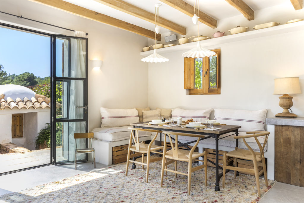 A cozy dining area with a wooden table and chairs, surrounded by white cushioned benches, exudes a sense of luxury travel. Pendant lights hang from a wooden beam ceiling. A small window and an open door reveal greenery outside, while a rug covers the floor. David Pearson Travel