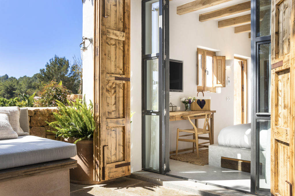 A cozy room with wooden accents offers a hint of luxury travel, featuring an open glass door leading outside. Inside, theres a bed, a wooden desk with a chair, and a small TV. Outside, a cushioned bench and potted plants are visible under the clear blue sky. David Pearson Travel