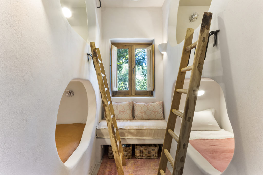 A cozy, small room features two built-in bunk beds with wooden ladders on either side. A central window lets in natural light, with a cushioned bench beneath it. Embrace minimalist luxury travel vibes with white walls and simple decor that exude warmth and comfort. David Pearson Travel