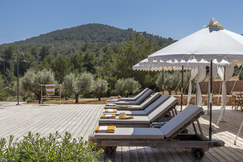 A scenic outdoor patio designed for luxury travel features several cushioned sun loungers facing a lush green hill. Each lounger is topped with a yellow towel. Nearby, a white canopy offers shade, while a tree-covered mountain rises majestically in the background under the clear blue sky. David Pearson Travel