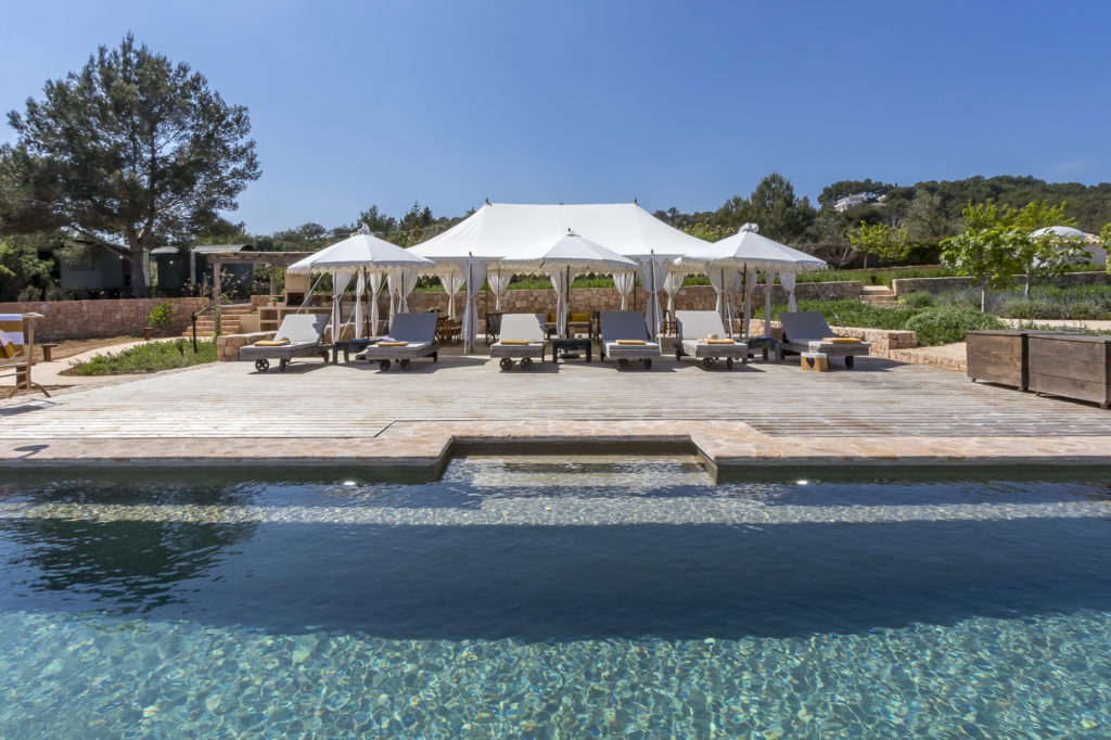 Poolside view with lounge chairs and white parasols on a wooden deck under a clear blue sky, perfect for a luxury travel retreat. The pool water reflects a pale turquoise color, surrounded by lush greenery and an enchanting landscape of trees. David Pearson Travel