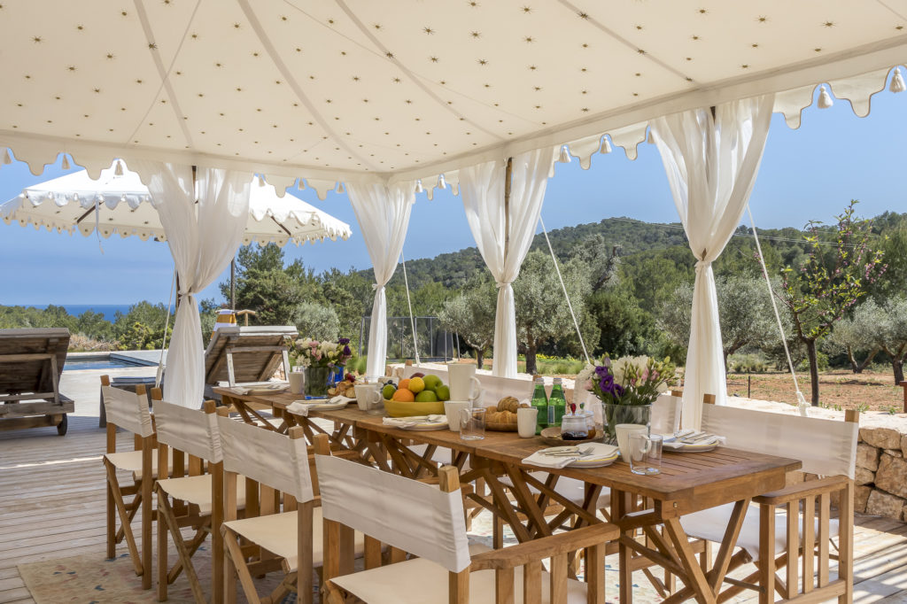 Experience luxury travel with an outdoor dining area under a white canopy, featuring a wooden table set with flowers, fruit, and beverages. In the background, loungers invite relaxation by the pool against a scenic backdrop of hills and trees. David Pearson Travel