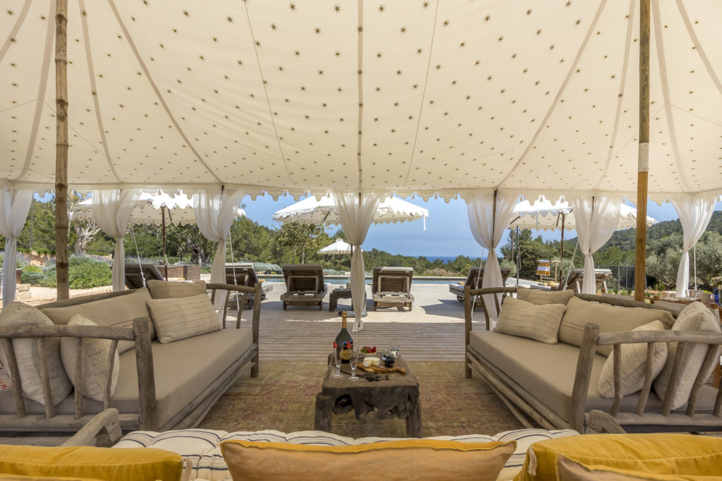 A luxurious outdoor lounge area with beige sofas under a grand decorative tent creates the perfect luxury travel experience. Sun loungers by the pool are framed by trees, offering a serene sea view. A small table elegantly displays wine and glasses, inviting indulgence in style. David Pearson Travel