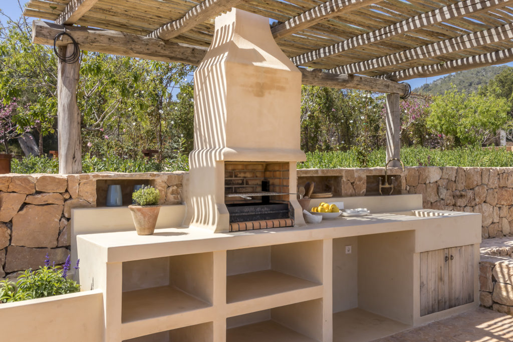 An outdoor kitchen with a beige stone barbecue grill under a wooden pergola evokes the essence of luxury travel. The countertop features open shelves, adorned with a potted plant and a bowl of lemons. Stone walls and lush greenery provide a stunning backdrop. David Pearson Travel