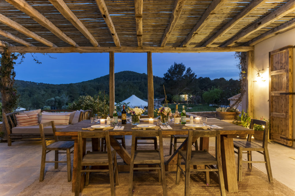 A rustic outdoor dining area exudes luxury travel vibes with a wooden table set elegantly with plates, glasses, and candles. Wooden chairs encircle the table under a pergola. In the background, a hilly landscape meets a tent. Evening light enhances the serene ambiance, perfect for indulgent escapes. David Pearson Travel