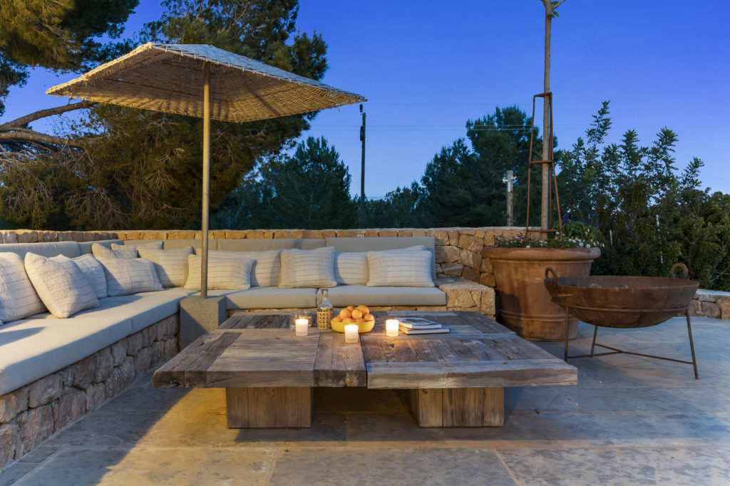 A cozy outdoor patio exudes luxury travel vibes with a rustic wooden table adorned with lit candles and fruit. Nestled under a woven umbrella is a large cushioned L-shaped sofa. Potted plants and a stone wall embrace the space, framed by trees and a clear blue evening sky. David Pearson Travel