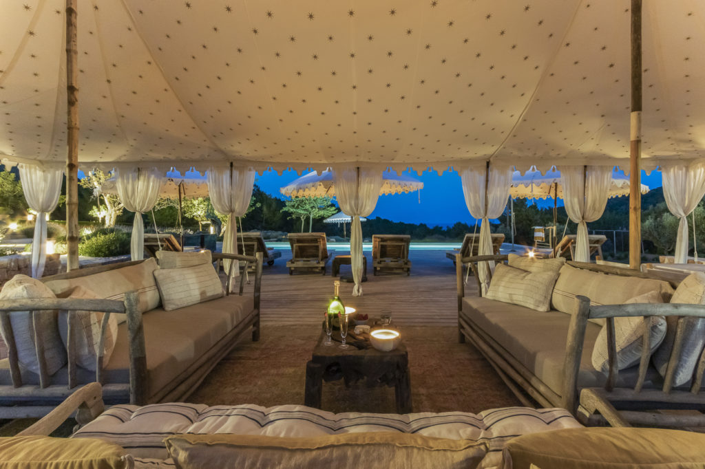 Experience luxury travel with a lavish outdoor lounge at night, featuring two sofas with cushions under a star-patterned canopy. Wooden tables hold lanterns and bowls, while draped curtains reveal lush greenery and a pool. Ambient lighting enhances the cozy atmosphere. David Pearson Travel