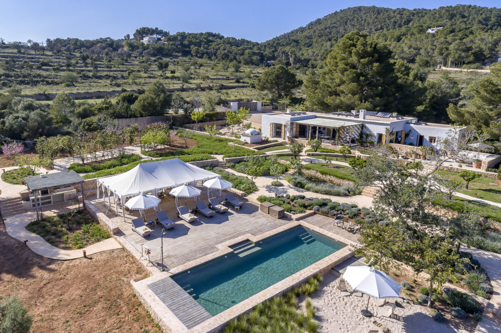 Aerial view of a luxurious villa, perfect for luxury travel enthusiasts, surrounded by greenery. It features a rectangular swimming pool, sun loungers under a white canopy, and lush gardens. The villa is set amidst hills and trees under a clear blue sky. David Pearson Travel