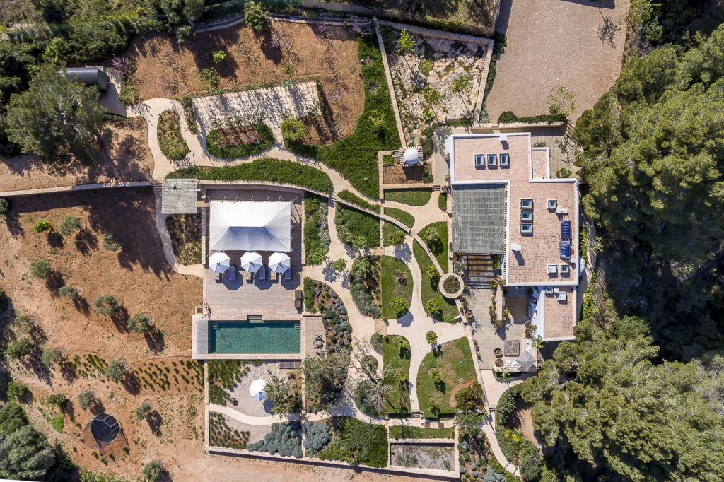 Aerial view of a landscaped property epitomizing luxury travel, featuring a rectangular swimming pool with sun loungers and umbrellas nearby, manicured gardens with circular paths, and a large building surrounded by trees and greenery. David Pearson Travel