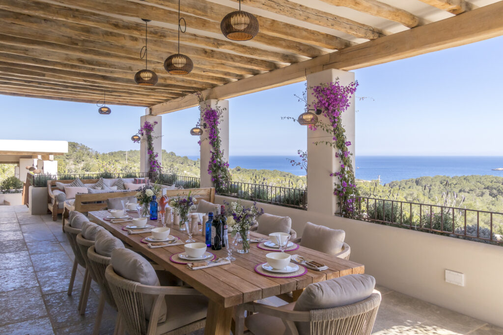 A spacious outdoor dining area epitomizes luxury travel with a long wooden table set elegantly with plates, glasses, and bottles. Cushioned chairs invite comfort while bougainvillea-adorned columns frame a breathtaking vista of lush landscapes and the shimmering ocean beyond. David Pearson Travel