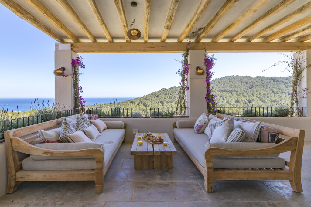 Experience luxury travel on this outdoor terrace, where two wooden couches adorned with cushions face each other. A small wooden table with snacks sits between them. The patio offers a breathtaking view of the ocean and lush greenery, enhanced by flowering vines climbing the columns. David Pearson Travel