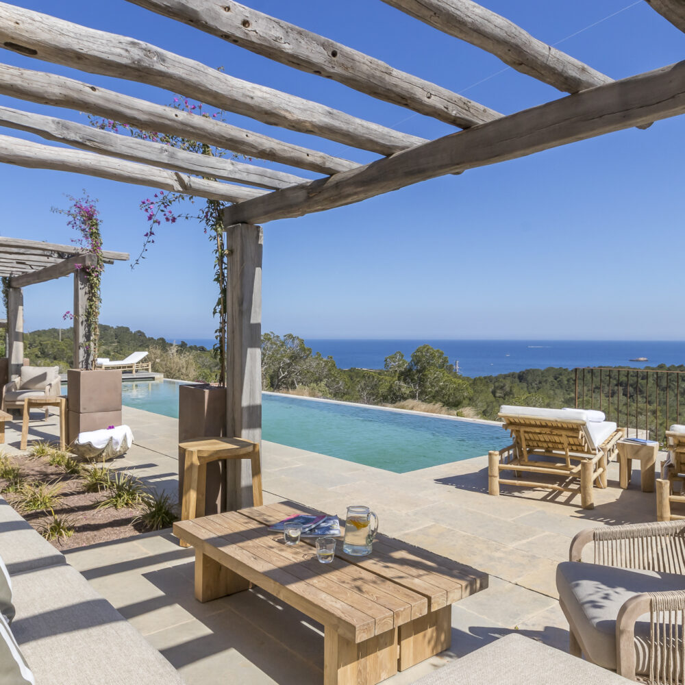 A luxurious outdoor patio with a pergola overlooks an infinity pool and a scenic ocean view, perfect for indulging in luxury travel. The area is furnished with comfortable seating and wooden lounge chairs. The sky is clear and blue, enhancing the serene atmosphere. David Pearson Travel