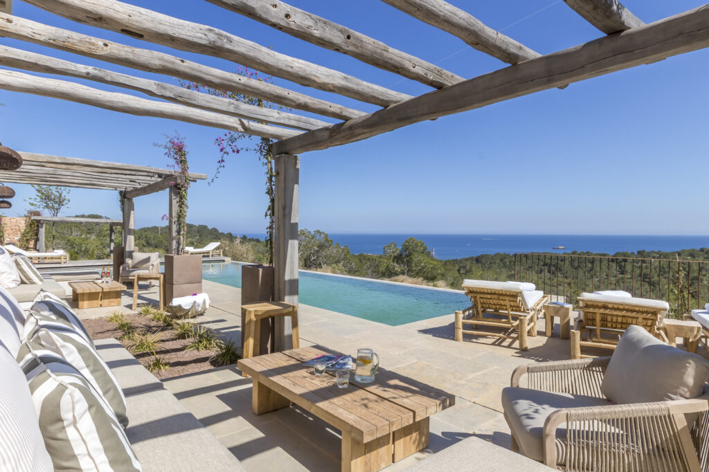 A luxurious outdoor patio with a pergola overlooks an infinity pool and a scenic ocean view, perfect for indulging in luxury travel. The area is furnished with comfortable seating and wooden lounge chairs. The sky is clear and blue, enhancing the serene atmosphere. David Pearson Travel