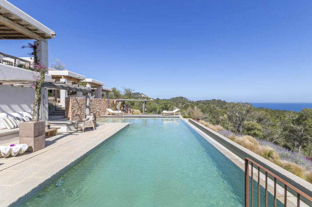 Experience the pinnacle of luxury travel at this villa with an infinity pool overlooking a breathtaking landscape. The modern architecture features white walls, wooden beams, and a sunbathing area with loungers. Mountains and a glimpse of the ocean are visible under a clear blue sky. David Pearson Travel