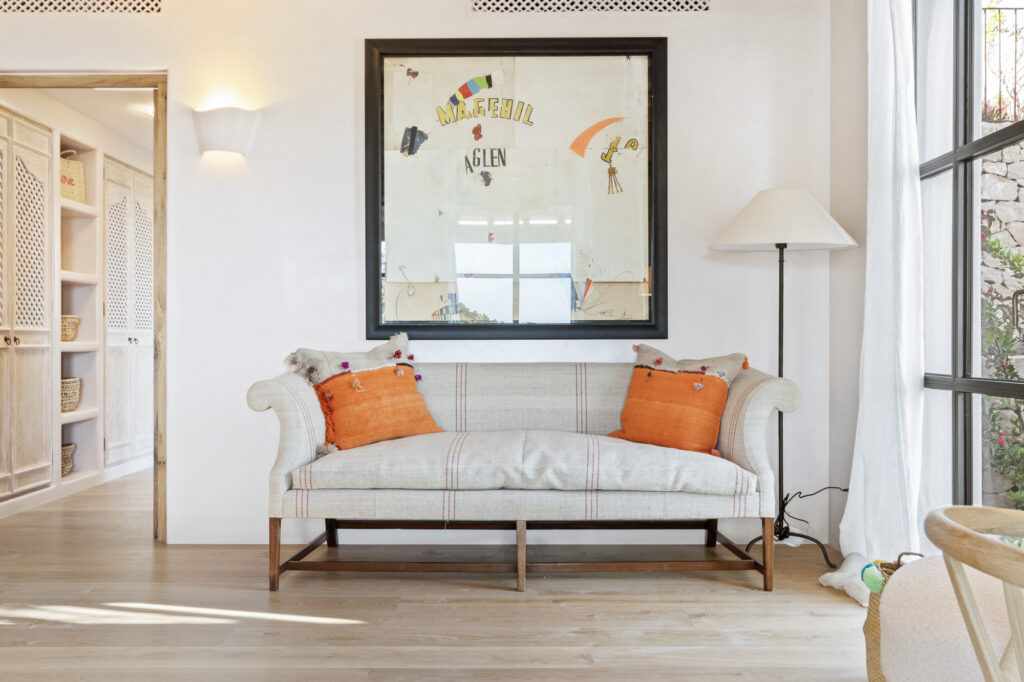 A cozy living room with a light-colored, checkered sofa adorned with orange pillows evokes the charm of luxury travel. Above the sofa hangs framed artwork, while a tall floor lamp stands beside a large window with sheer curtains. Built-in shelves are visible in an adjacent room. David Pearson Travel