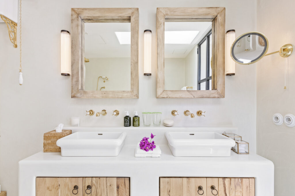 A modern bathroom exudes luxury travel vibes, with dual sinks and rectangular wooden mirrors above. Wall-mounted light fixtures and a round magnifying mirror add to the elegance. Purple flowers rest on a towel between the sinks, while toiletries are neatly arranged for a touch of sophistication. David Pearson Travel