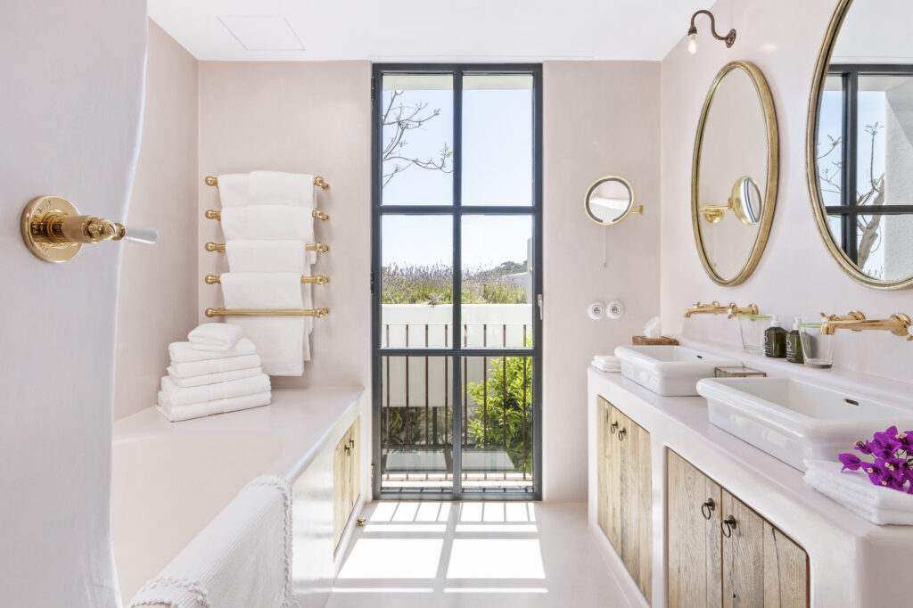 This bright bathroom evokes a sense of luxury travel with its large window, double sinks atop wooden cabinets, and two circular mirrors. A stack of white towels graces the bathtubs edge, while brass fixtures and a towel rack enhance the serene, airy space. David Pearson Travel
