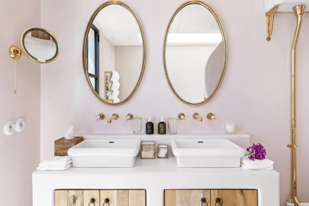 A bathroom with two oval mirrors above a double sink vanity exudes the elegance of luxury travel. The white sinks rest on wooden cabinets below, complemented by gold fixtures and a round magnifying mirror on the wall. Toiletries and folded towels are neatly arranged on the counter. David Pearson Travel