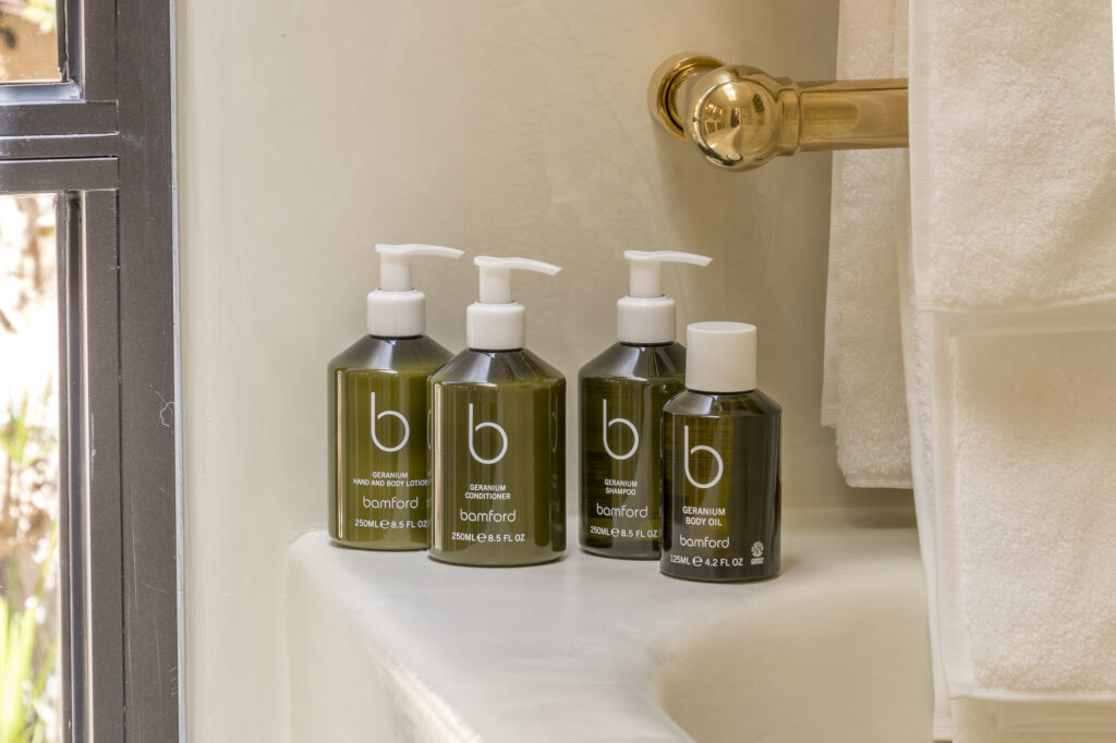 Four green bottles labeled bamford, perfect for luxury travel, with pump dispensers are lined up on a bathtub ledge. The bottles contain body wash, shampoo, conditioner, and body oil. A brass faucet and white towels are visible nearby. David Pearson Travel