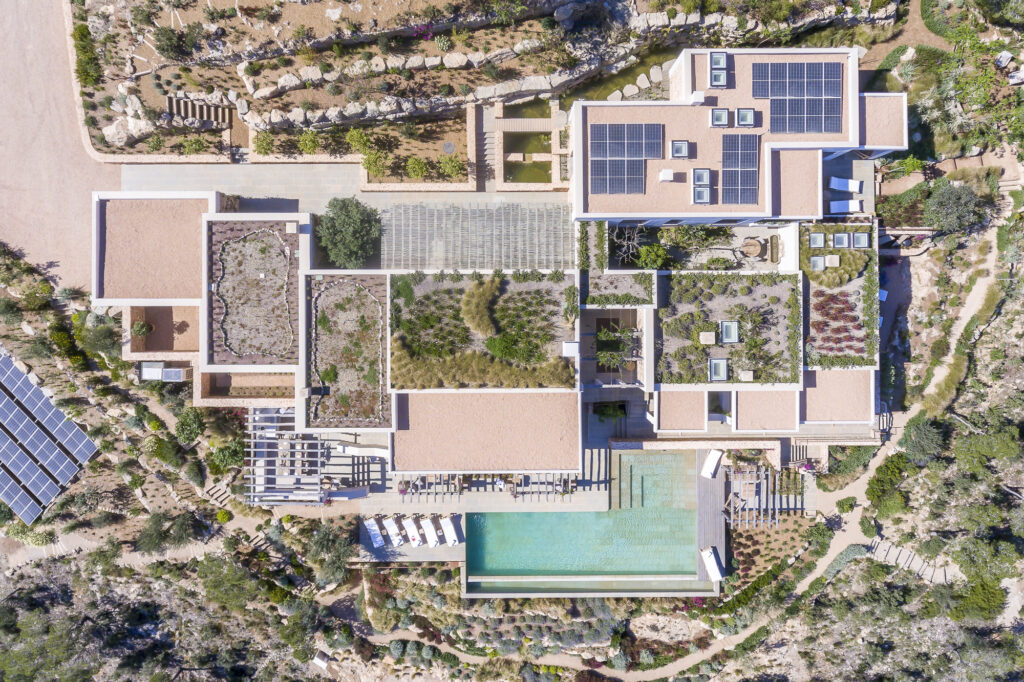 Aerial view of a modern luxury travel destination featuring interconnected structures with flat roofs, some adorned with solar panels. The property boasts landscaped gardens and a rectangular infinity pool, all set against the backdrop of arid, rocky terrain. David Pearson Travel