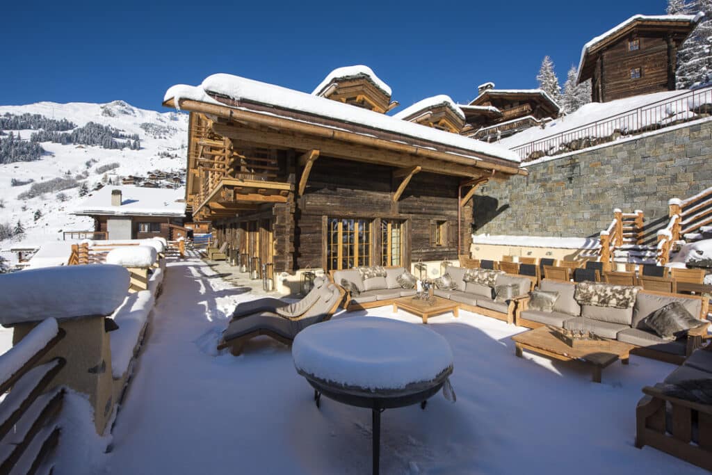 Snow-covered chalet with rustic wooden architecture and large patio, featuring cozy outdoor furniture. Perfect for luxury travel, the scene includes a circular snow-topped table set against a backdrop of snow-dusted mountains under a clear blue sky. David Pearson Travel