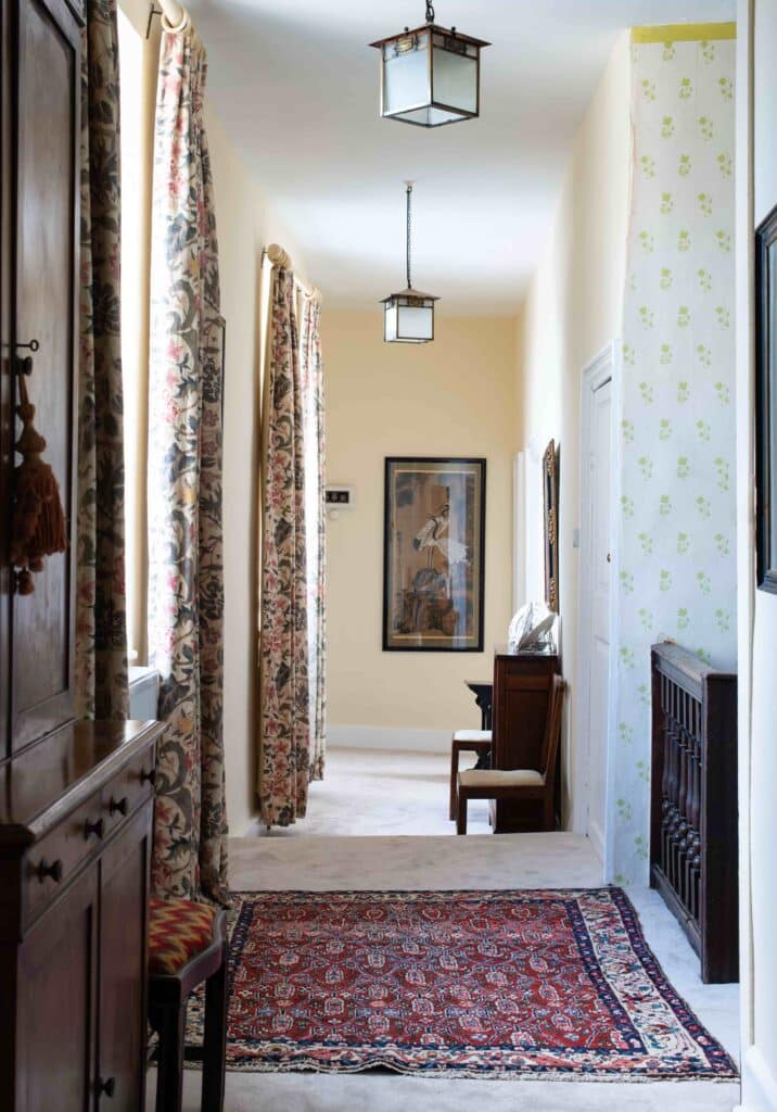 A narrow, sunlit hallway exudes the charm of luxury travel with its patterned curtains and decorative rug. Walls feature floral designs and artworks; a wooden cabinet and chair are elegantly positioned along the sides, while classic light fixtures hang from the ceiling. David Pearson Travel