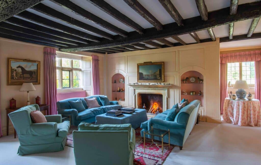 A cozy living room with a fireplace, surrounded by teal sofas and armchairs, adds a touch of luxury travel to your home. Natural light enters through large windows with pink curtains. The ceiling boasts exposed dark wooden beams, and artworks adorn the walls as a fire crackles warmly in the hearth. David Pearson Travel