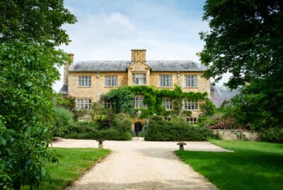 A large, picturesque stone house with ivy-covered walls stands at the end of a gravel path, offering a luxury travel experience. Surrounded by lush greenery and trees, the two-story building has multiple windows and a chimney, set under a partially cloudy sky. David Pearson Travel