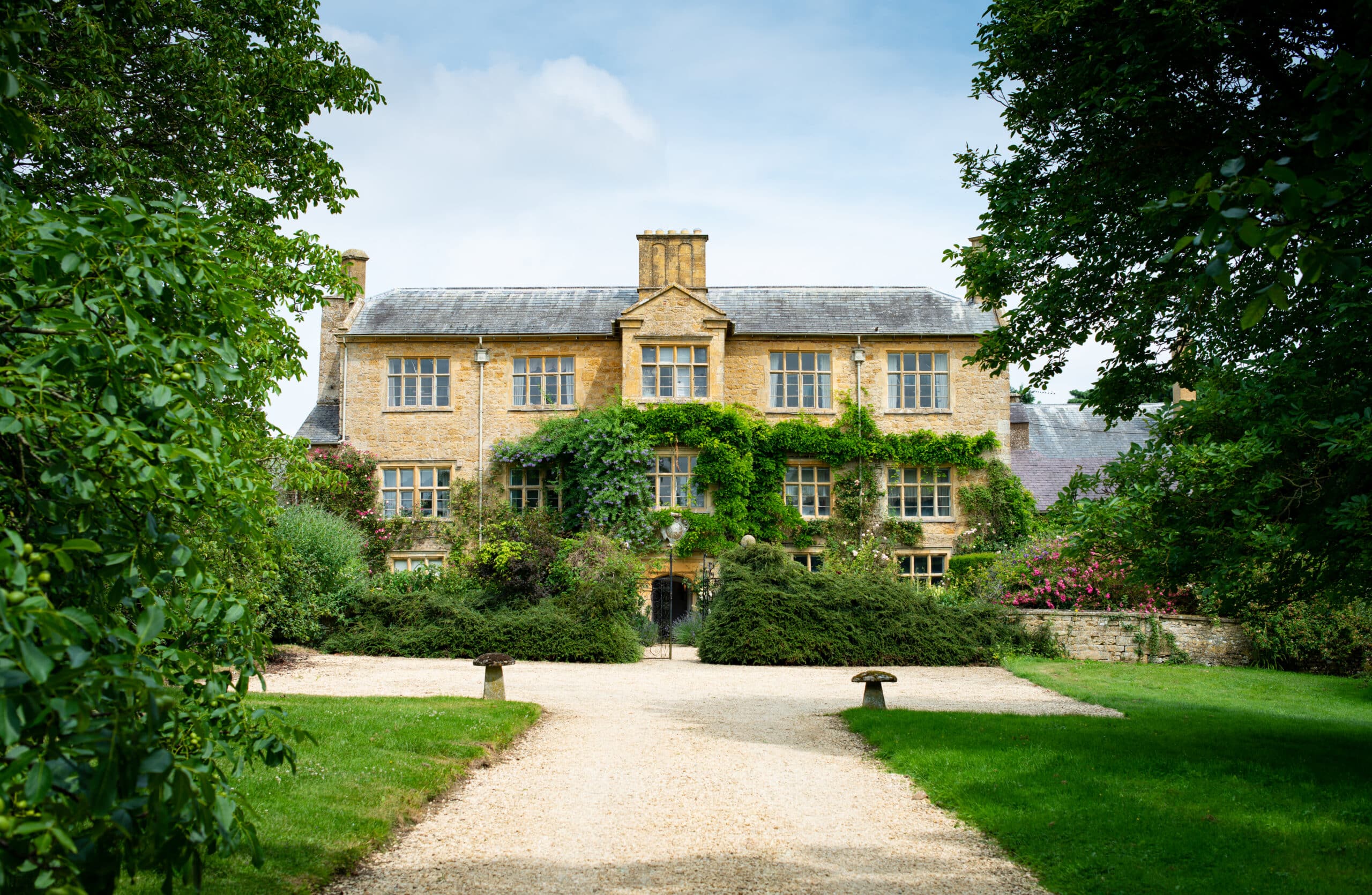 A large, picturesque stone house with ivy-covered walls stands at the end of a gravel path, offering a luxury travel experience. Surrounded by lush greenery and trees, the two-story building has multiple windows and a chimney, set under a partially cloudy sky. David Pearson Travel