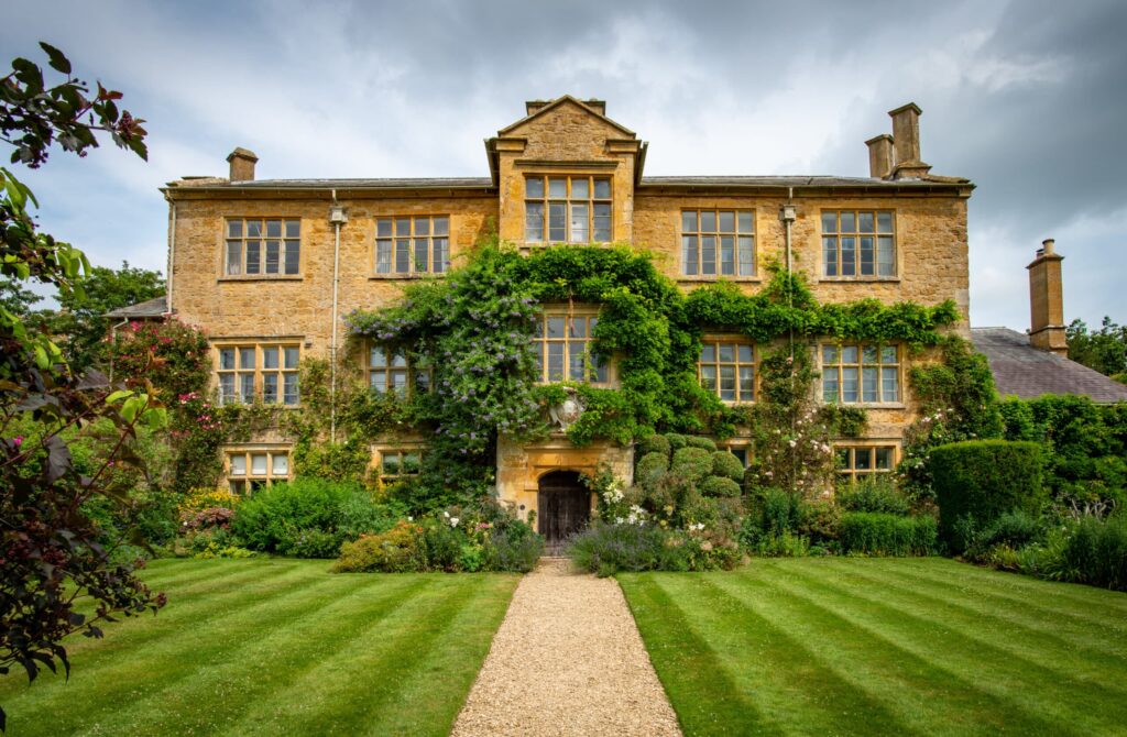 A large, historic stone house with many windows is covered in green ivy, exuding a sense of luxury travel. It has a central entrance and symmetrical gardens with a gravel path leading to the door. The lawn is neatly mowed, and the sky is overcast. David Pearson Travel
