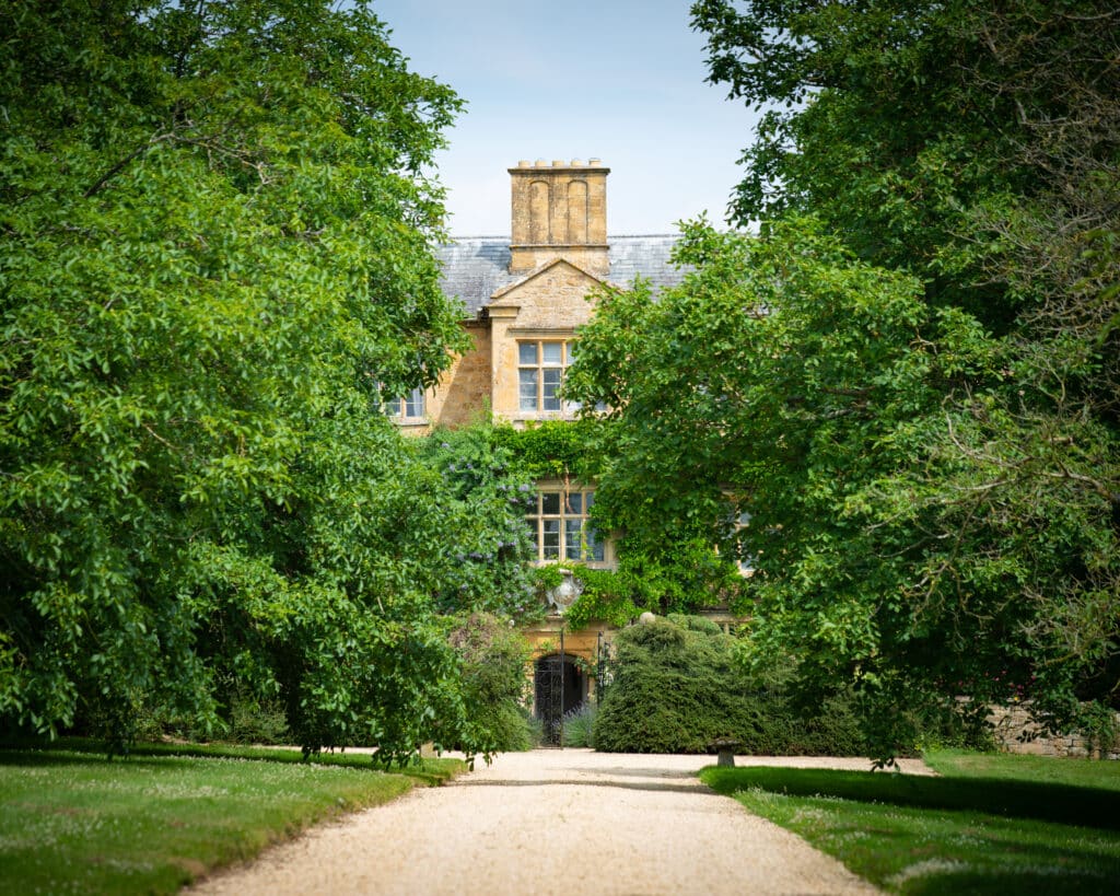 A picturesque stone house, perfect for luxury travel enthusiasts, is partially hidden by lush green trees and shrubs. A gravel pathway leads to the entrance, surrounded by manicured lawns. The house features a chimney and ivy-covered walls, exuding a charming countryside feel. David Pearson Travel