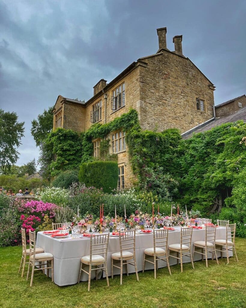An elegant outdoor dining setup exuding luxury travel vibes features a long table adorned with flowers and red napkins. White chairs surround the table on a lush green lawn, set against a backdrop of an ivy-covered brick building under a cloudy sky. David Pearson Travel