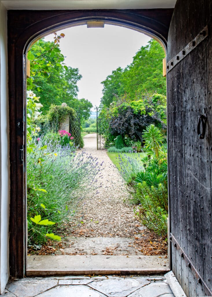 View through an open wooden door revealing a lush garden path, reminiscent of a luxury travel destination, lined with lavender and greenery, leading to a distant gate. Trees and dense foliage surround the serene scene in the background under a cloudy sky. David Pearson Travel