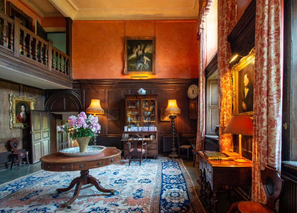 A vintage room exuding luxury travel vibes features ornate wooden furniture and a tall ceiling. A large round table with a floral centerpiece rests on a patterned carpet. The walls boast paintings, while lamps cast a warm glow. A balcony and expansive windows complete the elegant ambiance. David Pearson Travel